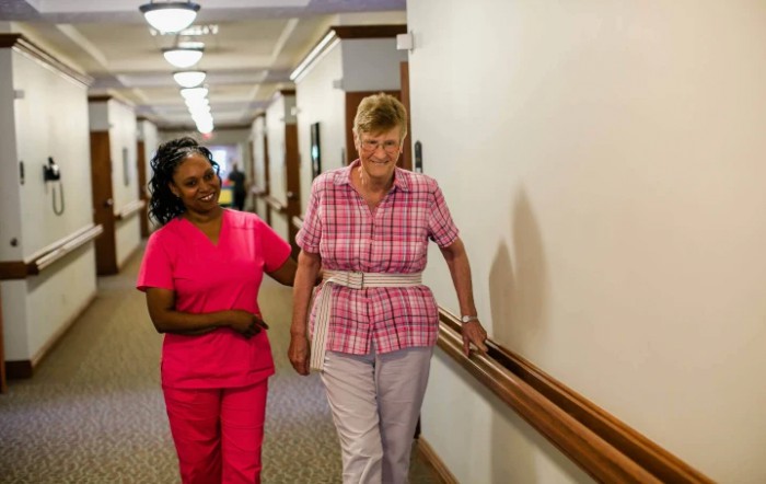 A healthcare worker assists an elderly woman walking down a corridor.