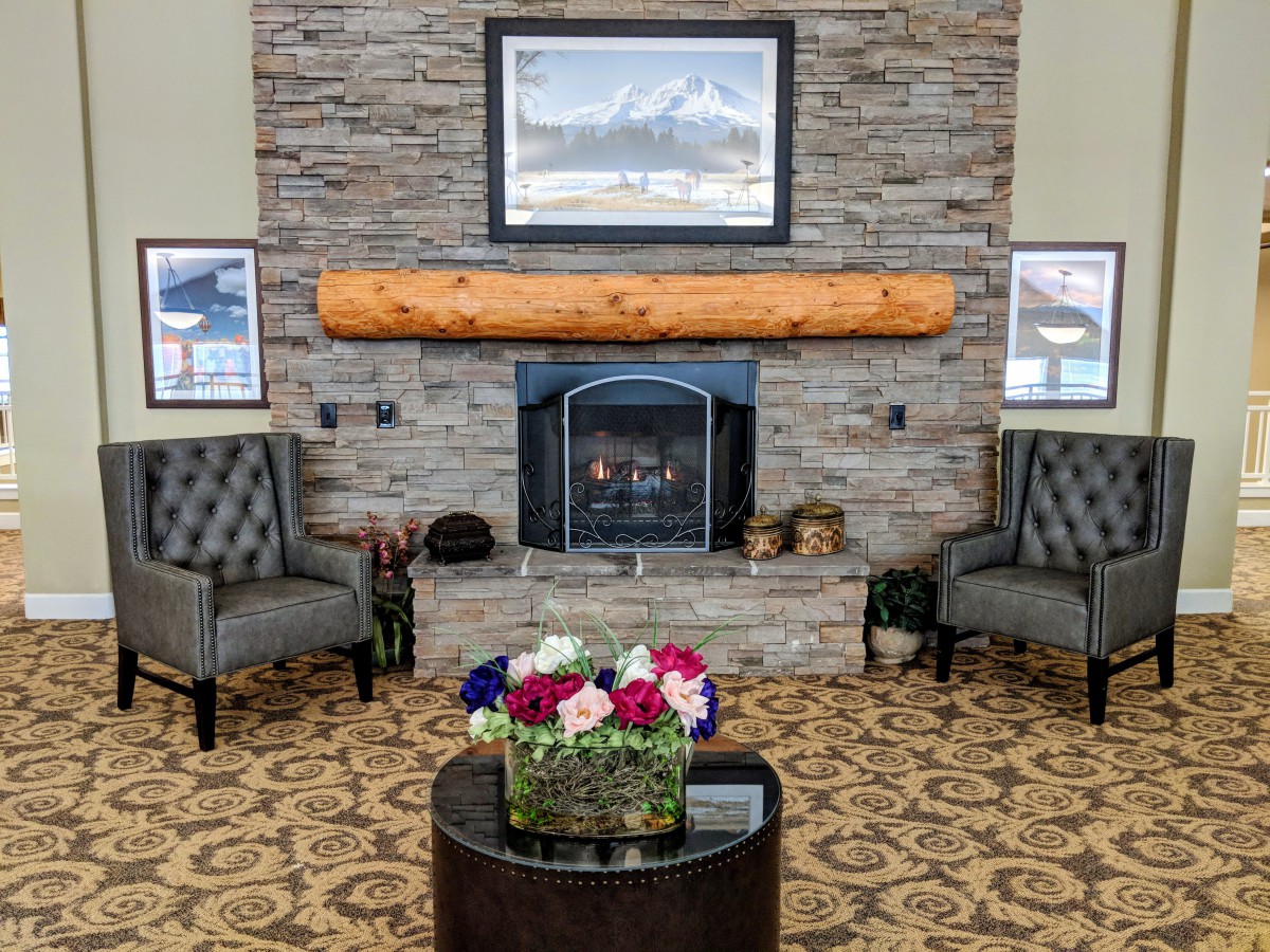 A cozy lounge area featuring a stone fireplace with two chairs, a floral centerpiece on a round table, and mountain artwork above the mantel.