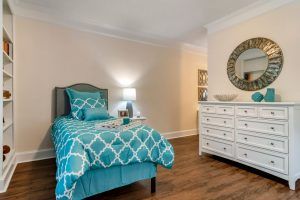 A cozy bedroom with a single bed featuring blue patterned bedding, a white dresser with a large mirror above it, and warm wooden flooring.