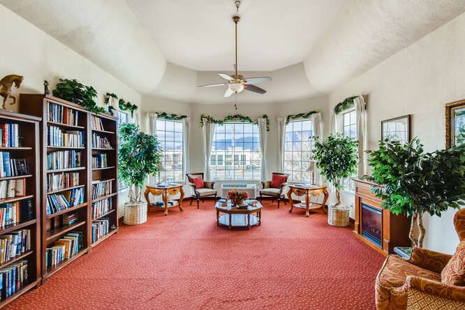 A cozy living room with large windows, bookshelves, and a seating area surrounded by plants.