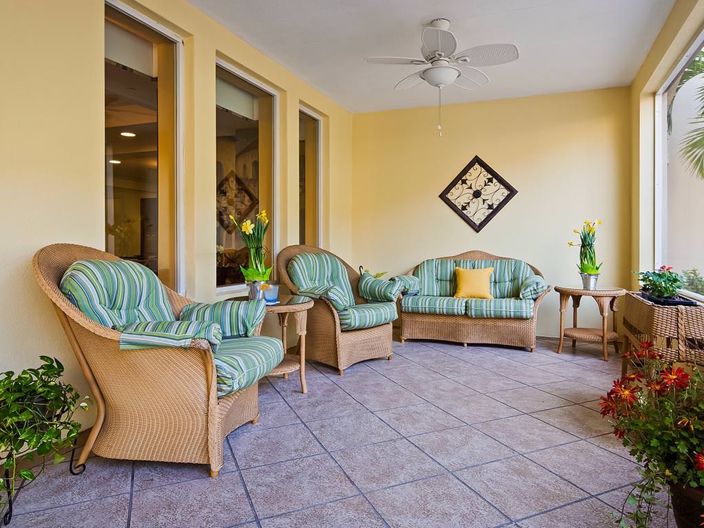 A cozy living room with rattan furniture and vibrant cushions, featuring large windows and decorative plants.