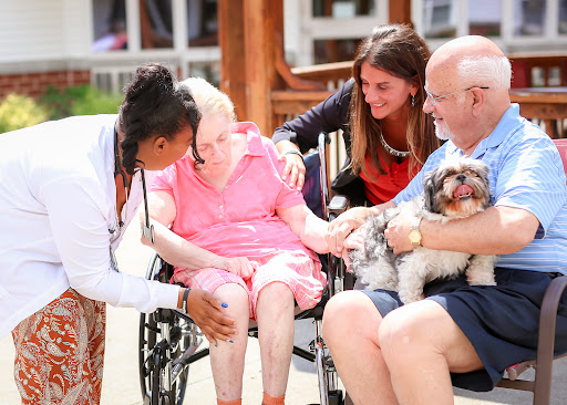 A caregiver and family members providing companionship and support to an elderly woman in a wheelchair, enjoying a sunny day with a pet.