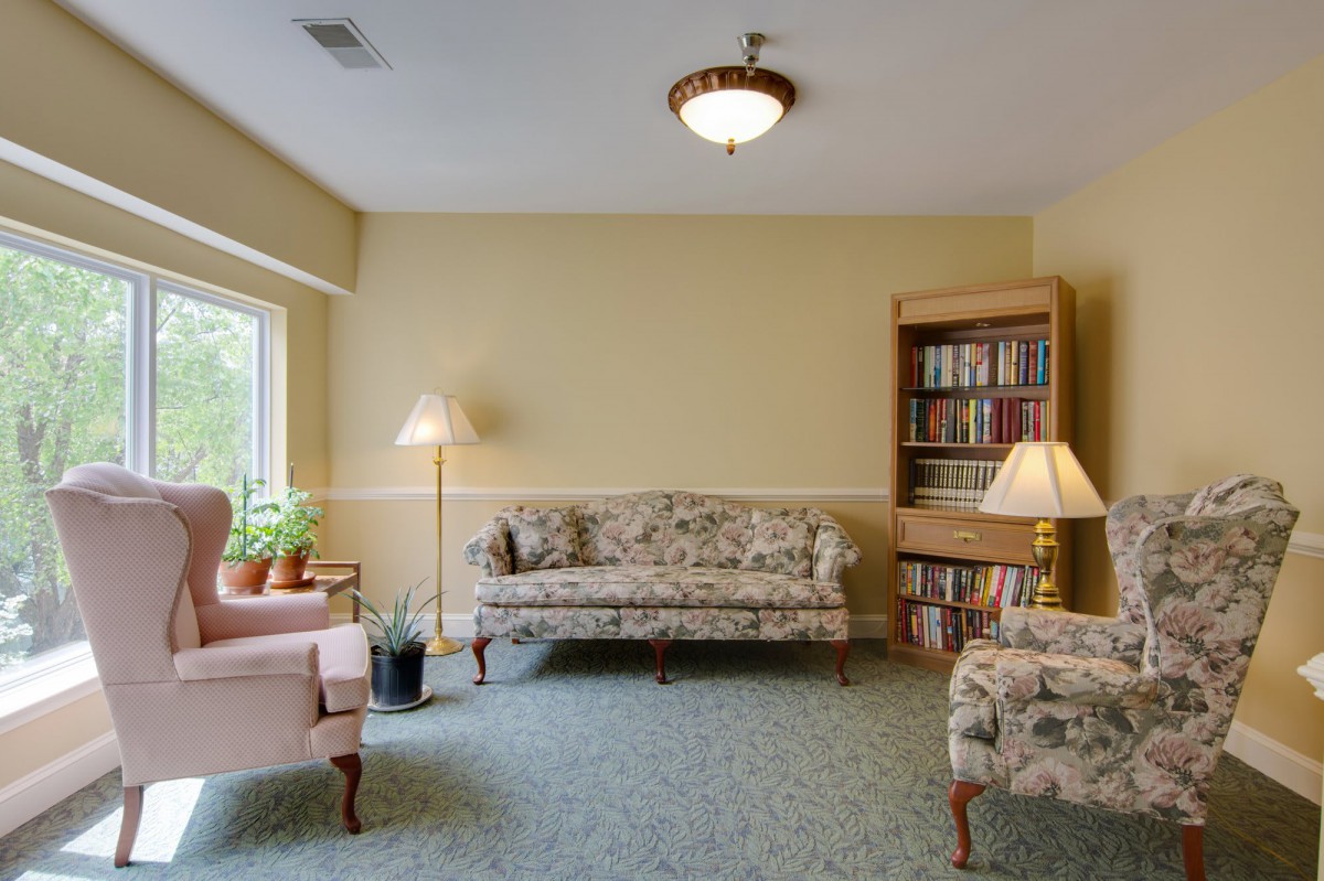 A cozy living room with a floral sofa, armchairs, bookshelves, and warm lighting.