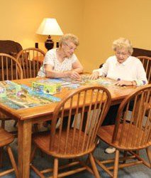 Two elderly women working on a puzzle together at a wooden table.