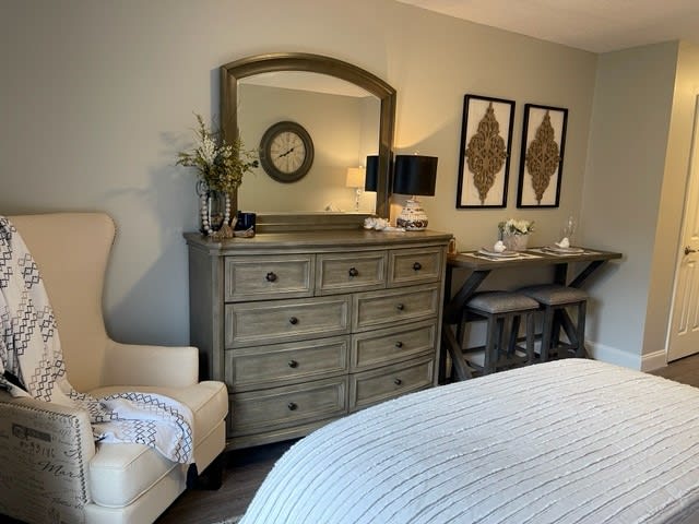 A cozy bedroom featuring a dresser with a mirror, decorated with a plant and framed artwork, alongside a comfortable chair and neatly made bed.