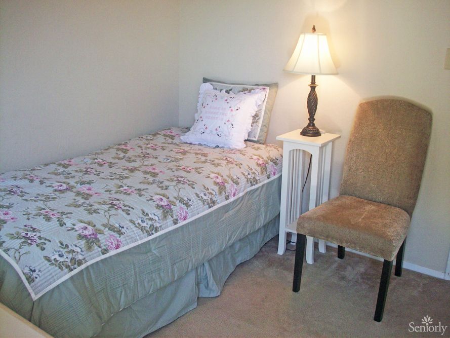 A cozy bedroom with a single bed, nightstand, and chair under a warm lamp.