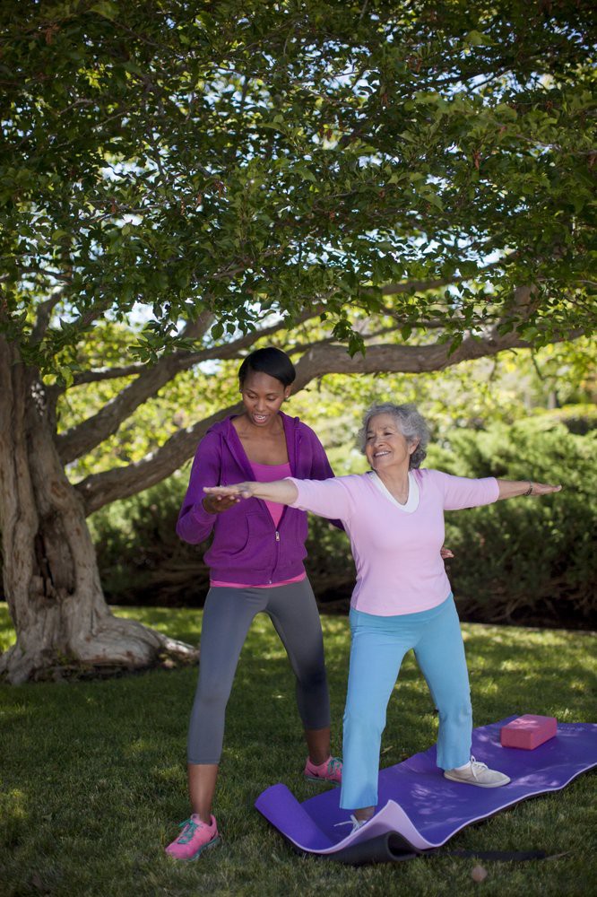 A yoga instructor guides an elderly woman in a yoga pose under a tree.