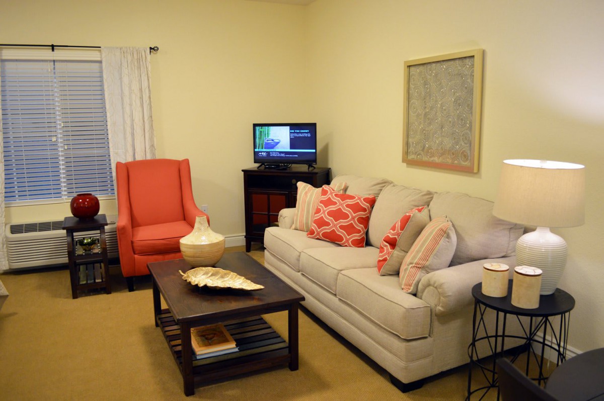 A cozy living room with a beige sofa, accented by red pillows and an armchair, centered around a wooden coffee table and a small television.