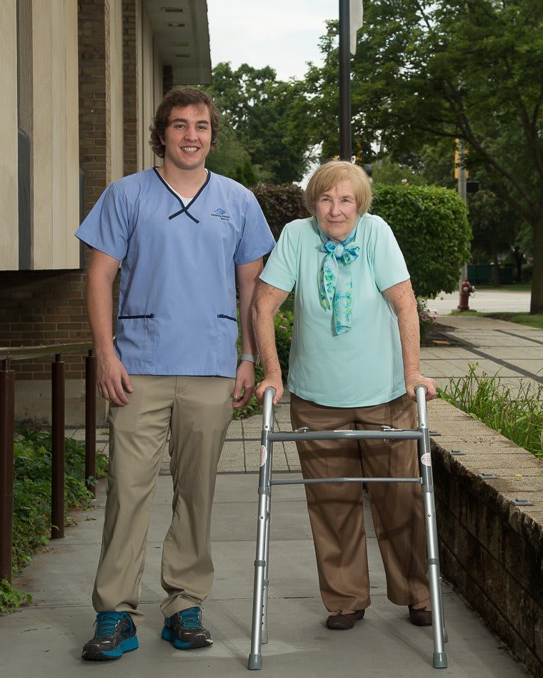 A caregiver assisting an elderly woman using a walker outside.