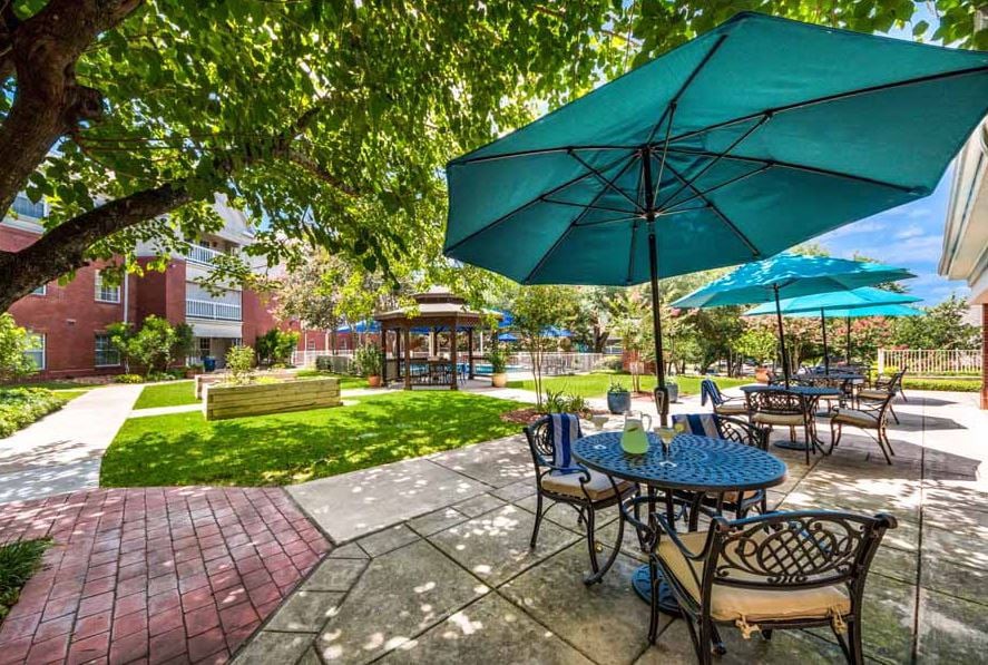 A charming outdoor seating area with tables and blue umbrellas, surrounded by a lush garden and gazebo under the shade of trees.