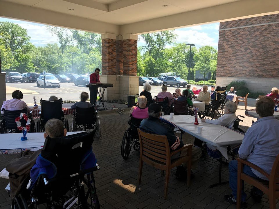 An outdoor musical performance attended by an audience, including elderly individuals in wheelchairs.