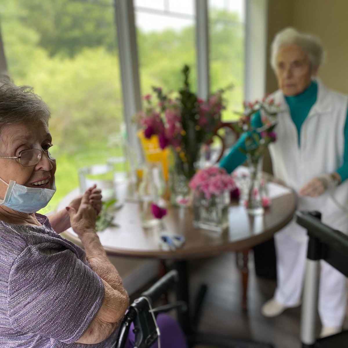 Two elderly women enjoy a social moment by a window with flowers on the table.