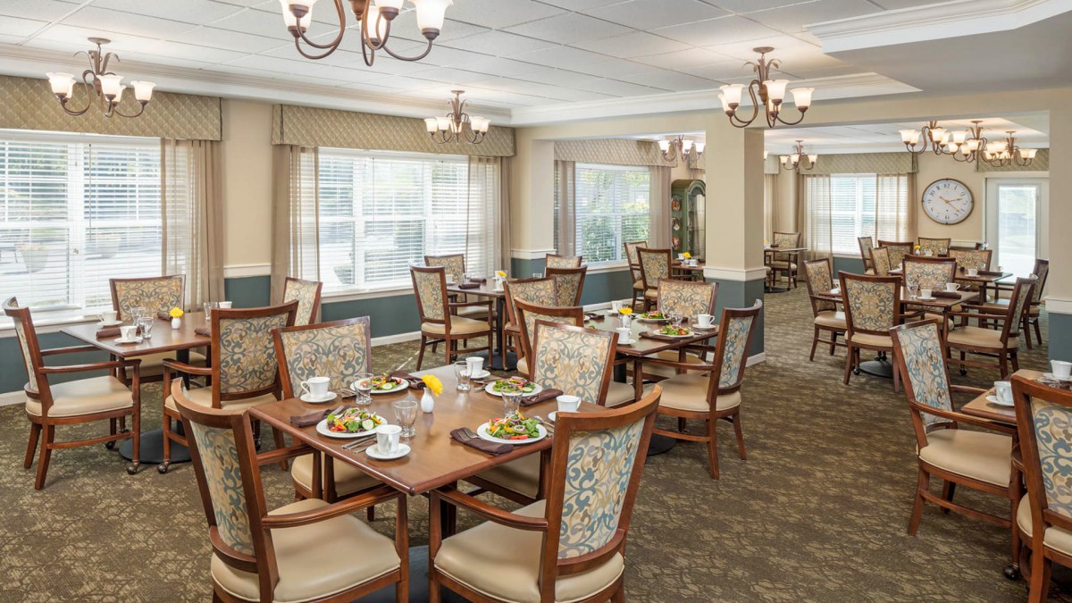A well-lit dining area with elegantly set tables and chairs, featuring large windows and chandeliers.