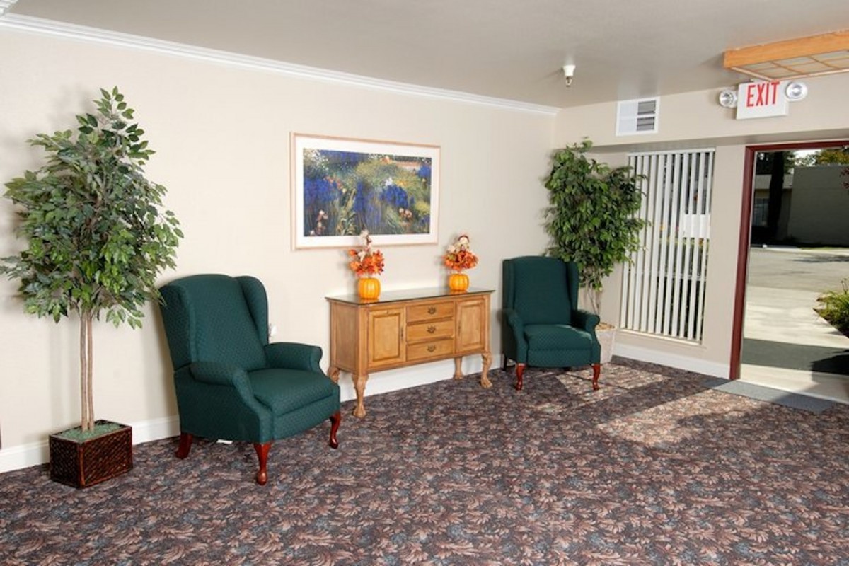 A neatly arranged lobby area with two green upholstered chairs, a wooden console table with decorative vases, framed artwork on the wall, and potted plants in the corners.