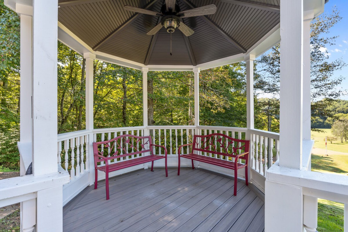 A peaceful gazebo with red benches and a ceiling fan, surrounded by trees.