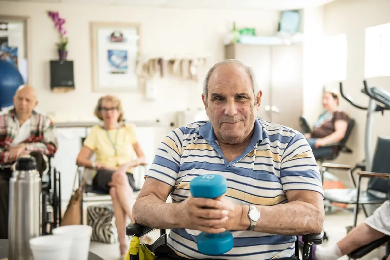 An elderly man holding a dumbbell while seated, with another person in the background, in an indoor setting.