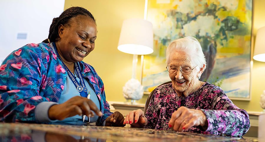 Two women, one elderly and one caregiver, happily work on a puzzle together indoors.