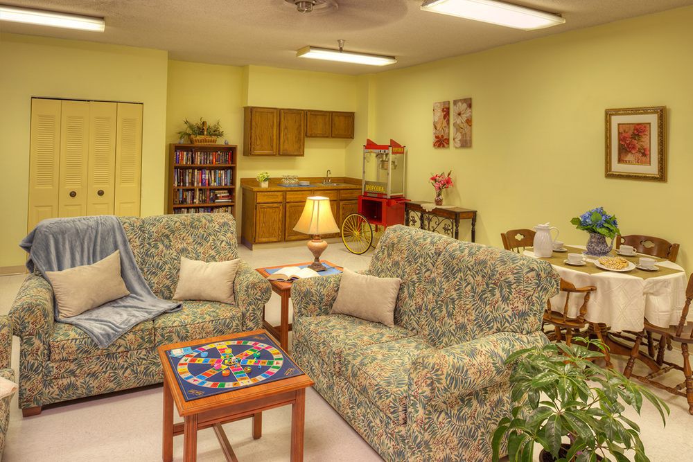 A cozy living room with floral-patterned sofas, a small table, and a lamp, adjacent to a kitchenette and bookshelf.