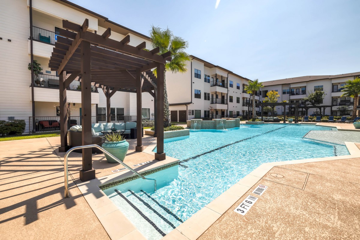 A modern apartment complex features a large outdoor swimming pool with palm trees and a pergola under a sunny sky.