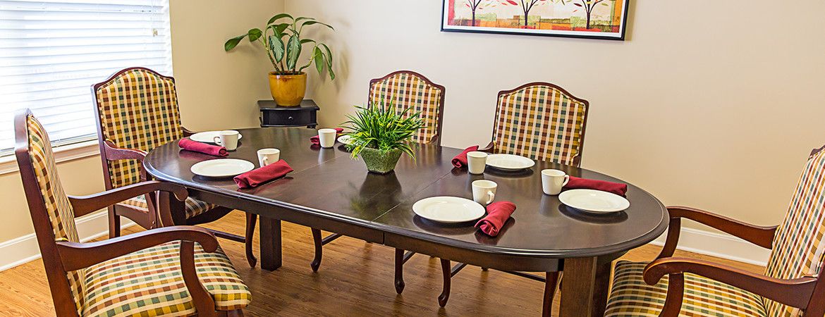 A dining room table set with plates, cups, and red napkins, surrounded by chairs and decorated with a potted plant.