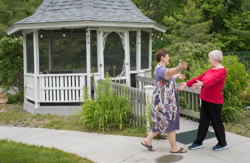 Two people greeting each other with a hug near a gazebo in a garden setting.