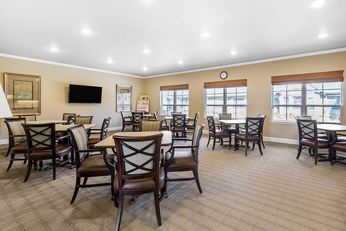 A spacious dining area with multiple tables and chairs, featuring large windows and a wall-mounted television.