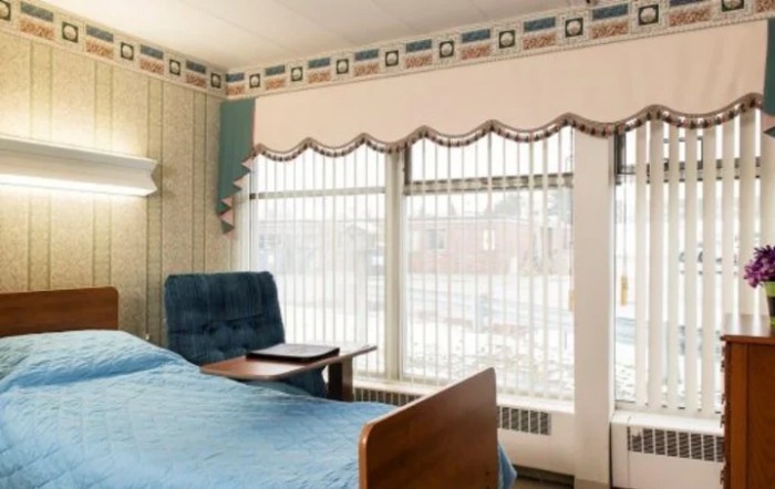 A cozy room with a bed, chair, and large windows adorned with blinds and curtains.