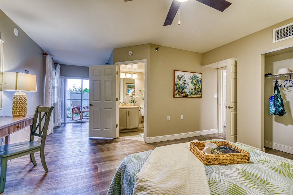 A cozy bedroom with a stylish interior, featuring a comfortable bed, ceiling fan, and wooden flooring. An open door leads to the bathroom, and artwork adorns the wall.