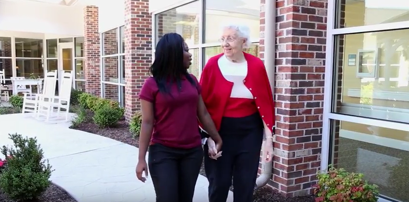 A caregiver and an elderly woman walking together outside a building.