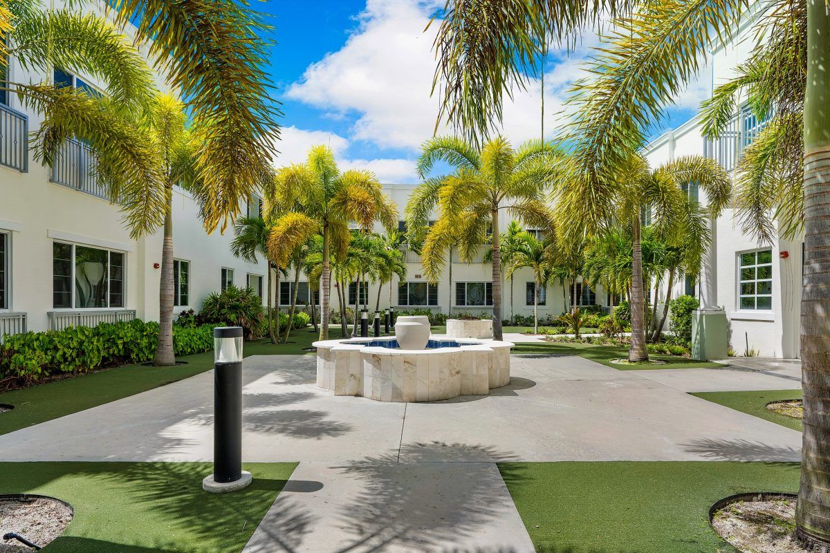A serene courtyard featuring palm trees and a central fountain under a sunny sky.
