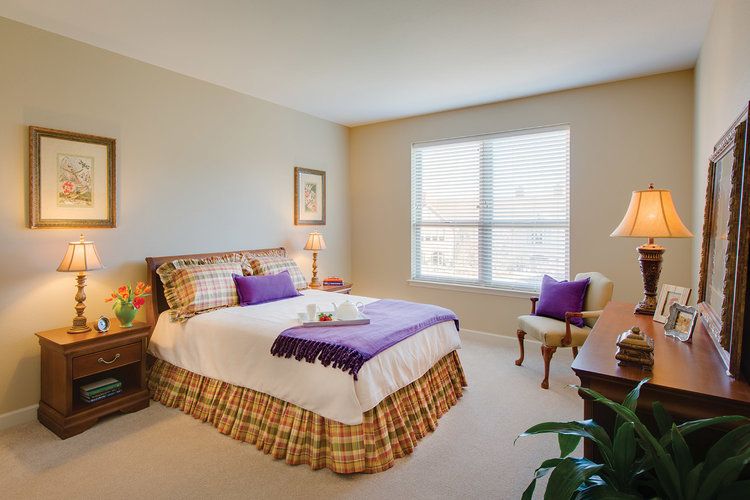A cozy bedroom with a neatly made bed featuring purple accents and a large window letting in natural light.