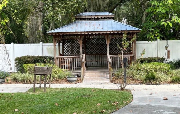 A wooden gazebo in a garden setting, surrounded by greenery and a walkway.