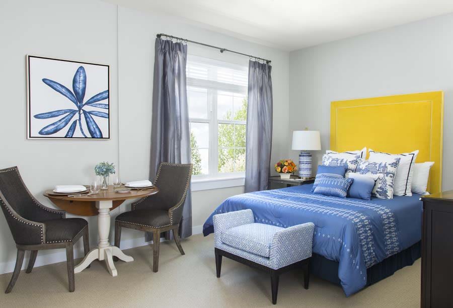 A stylish bedroom with blue bedding and a yellow headboard, featuring a small dining area by the window.
