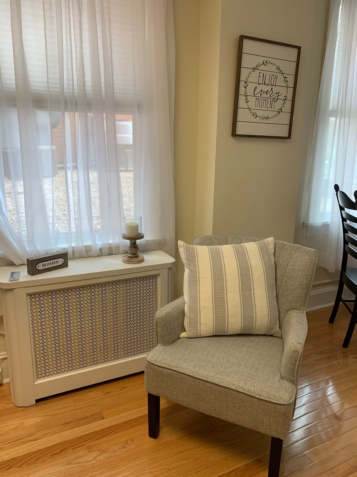 A cozy living room corner with a striped pillow on a chair next to a window and radiator cover.