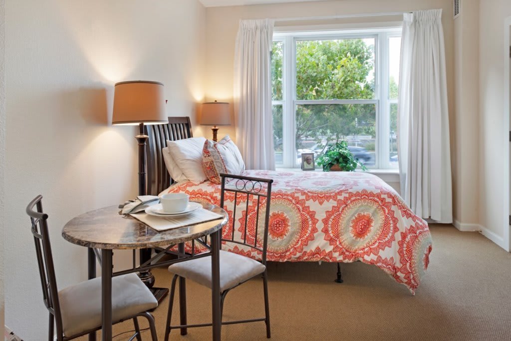 A modern bedroom with a colorful bedspread, a small dining table with two chairs, and large windows allowing natural light.