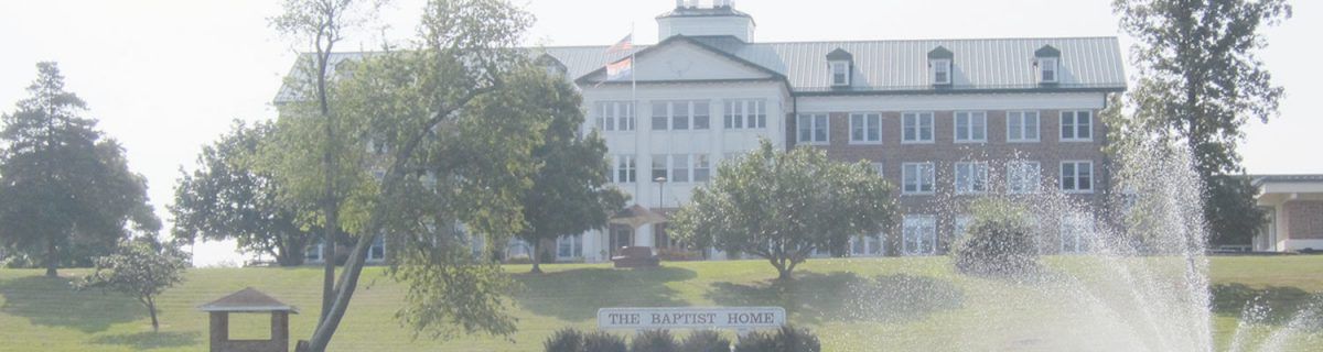 A large building with a sign reading 'The Baptist Home', surrounded by trees and an American flag.