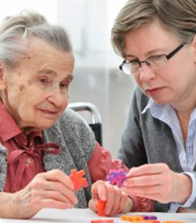 An elderly woman receiving assistance with a puzzle from a caregiver.