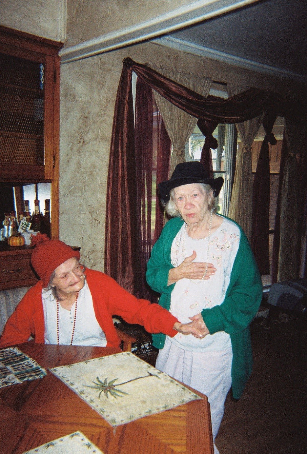 Two elderly women dressed in costumes, with one sitting and the other standing, sharing a light moment indoors.