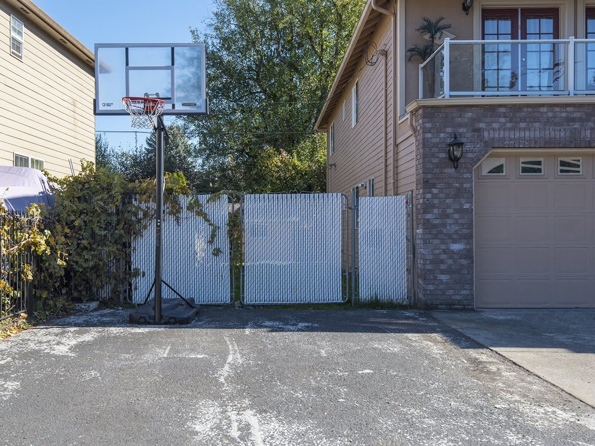 A driveway with a basketball hoop next to a house and a white fence.