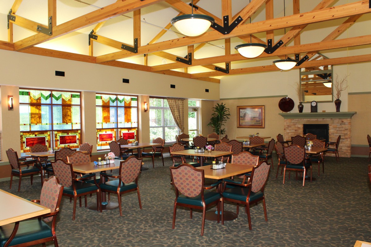 A spacious and well-lit restaurant interior with multiple tables and chairs, featuring decorative windows and overhead ceiling beams.