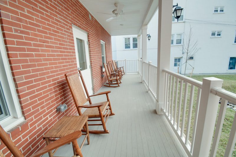 A spacious porch with wooden rocking chairs and a white railing, set against a brick wall.