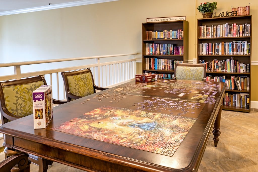A cozy library setting featuring a wooden table ready for a board game session, surrounded by bookshelves filled with various books.