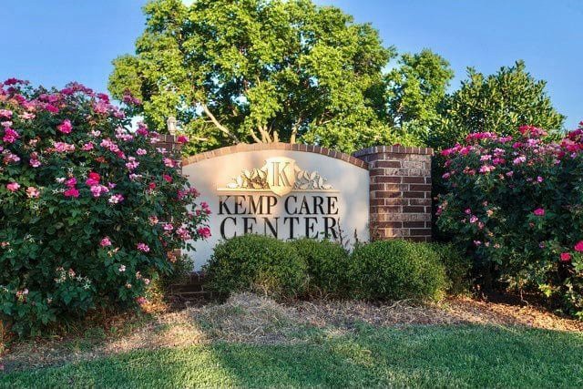 A sign for Kemp Care Center surrounded by vibrant flowers and lush greenery.