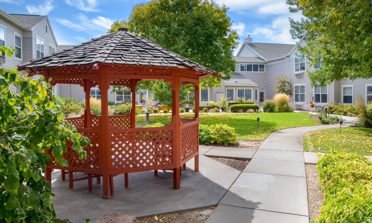 A charming wooden gazebo in a beautifully landscaped garden with a pathway leading to a large house.