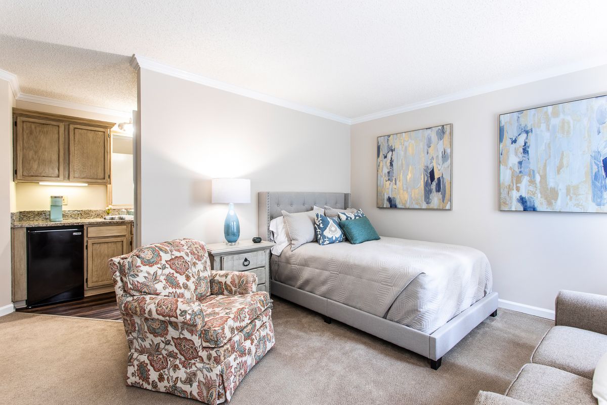 A cozy bedroom featuring a neatly made bed, floral armchair, and decorative artwork.