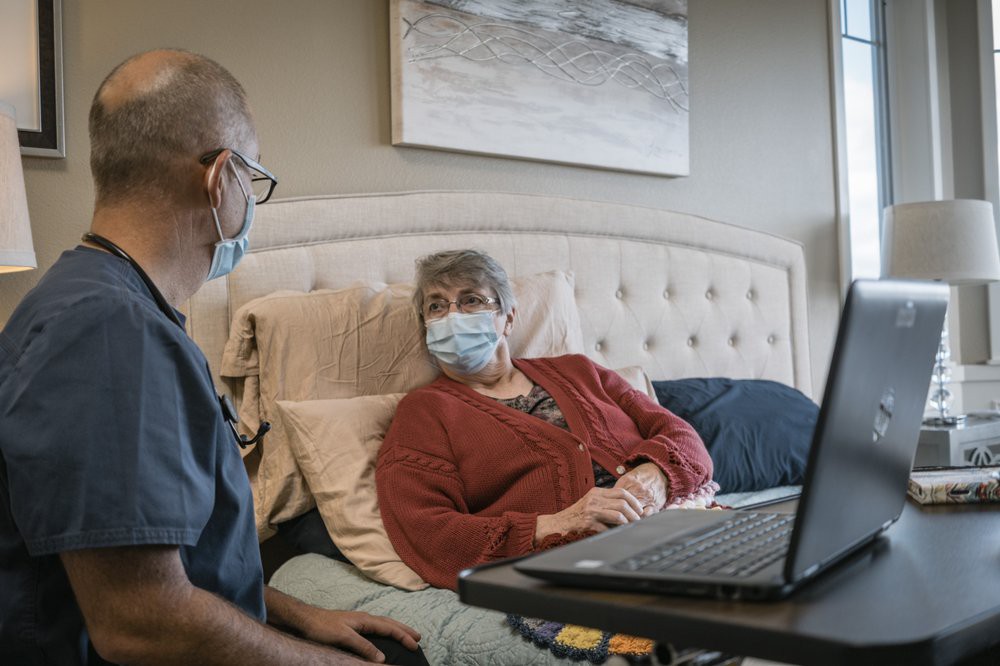A healthcare worker visits a patient at home, both wearing masks, and engages in conversation with the help of a laptop.