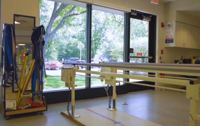 A physical therapy room equipped with parallel bars overlooking a window with a view of trees outside.