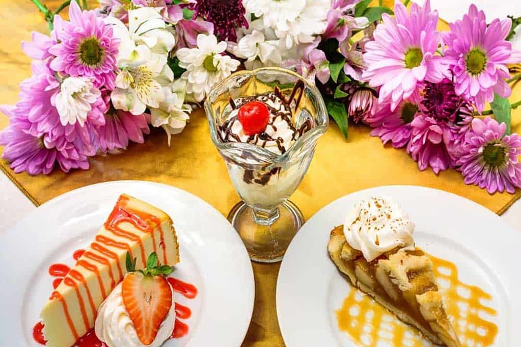 A delightful assortment of desserts featuring a sundae with a cherry on top, surrounded by slices of strawberry cake and banana cake, garnished with colorful flowers.
