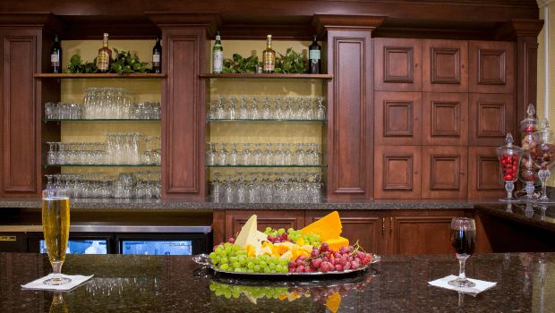 A kitchen or bar setup featuring a tray of assorted fruits on a granite countertop with wine glasses and wooden cabinets in the background.