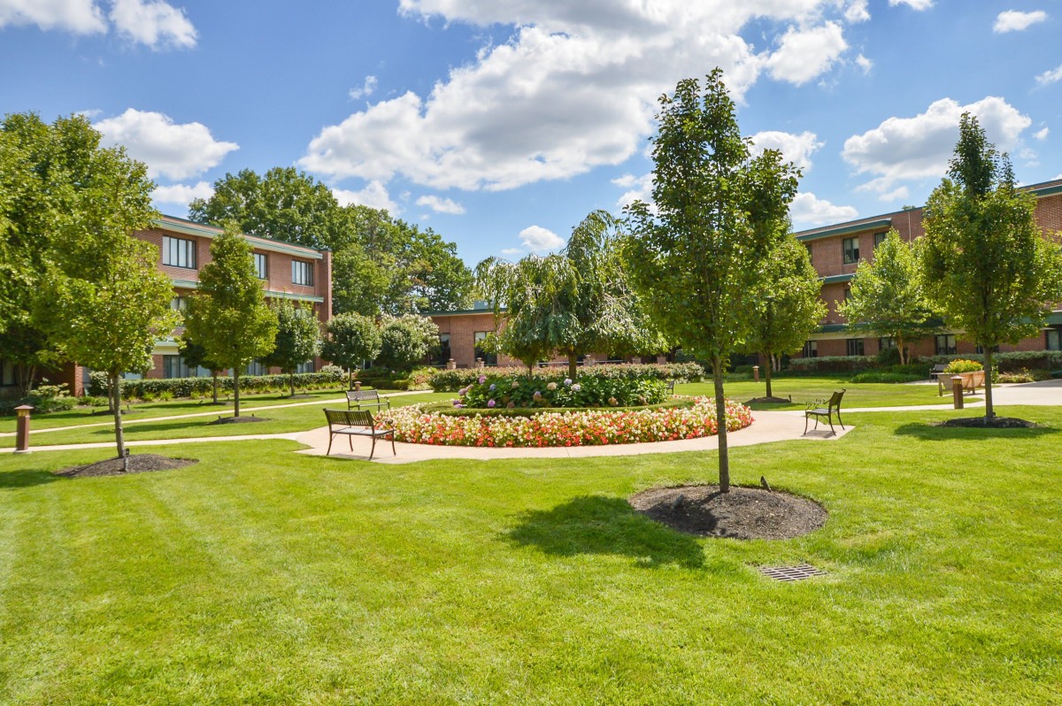 A beautifully landscaped garden area with trees, benches, and surrounding buildings under a partly cloudy sky.
